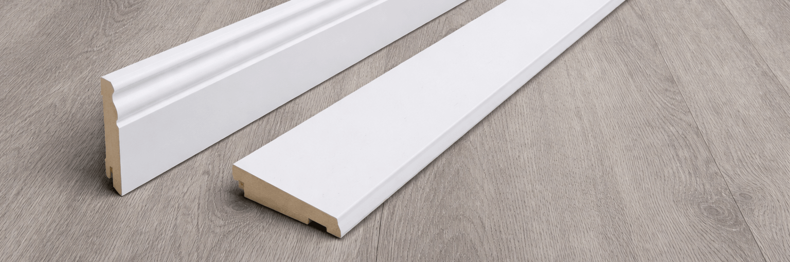 Junge Frau in Latzhose kniet auf dem Boden und hält lächelnd ein helles Laminatbrett in einem renovierungsbedürftigen Raum