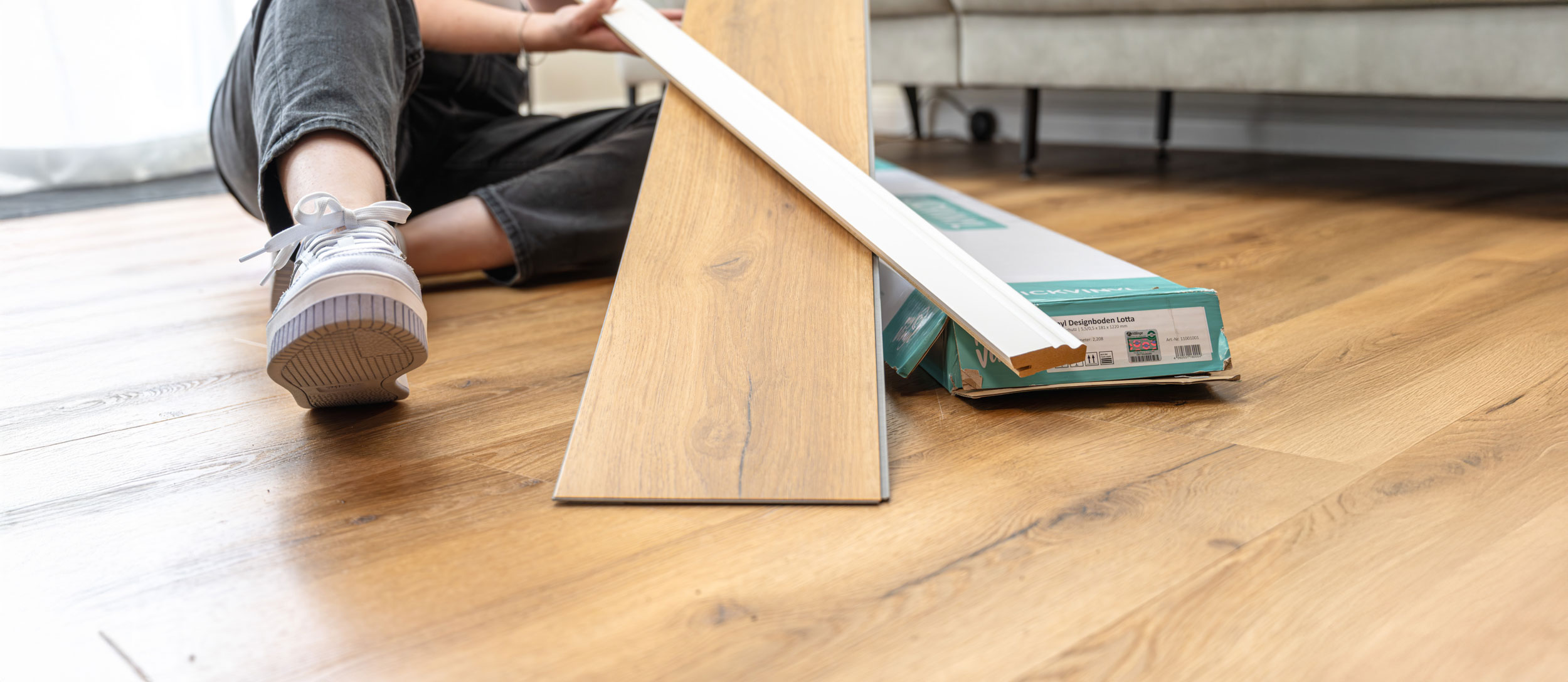 Junge Frau in Latzhose kniet auf dem Boden und hält lächelnd ein helles Laminatbrett in einem renovierungsbedürftigen Raum