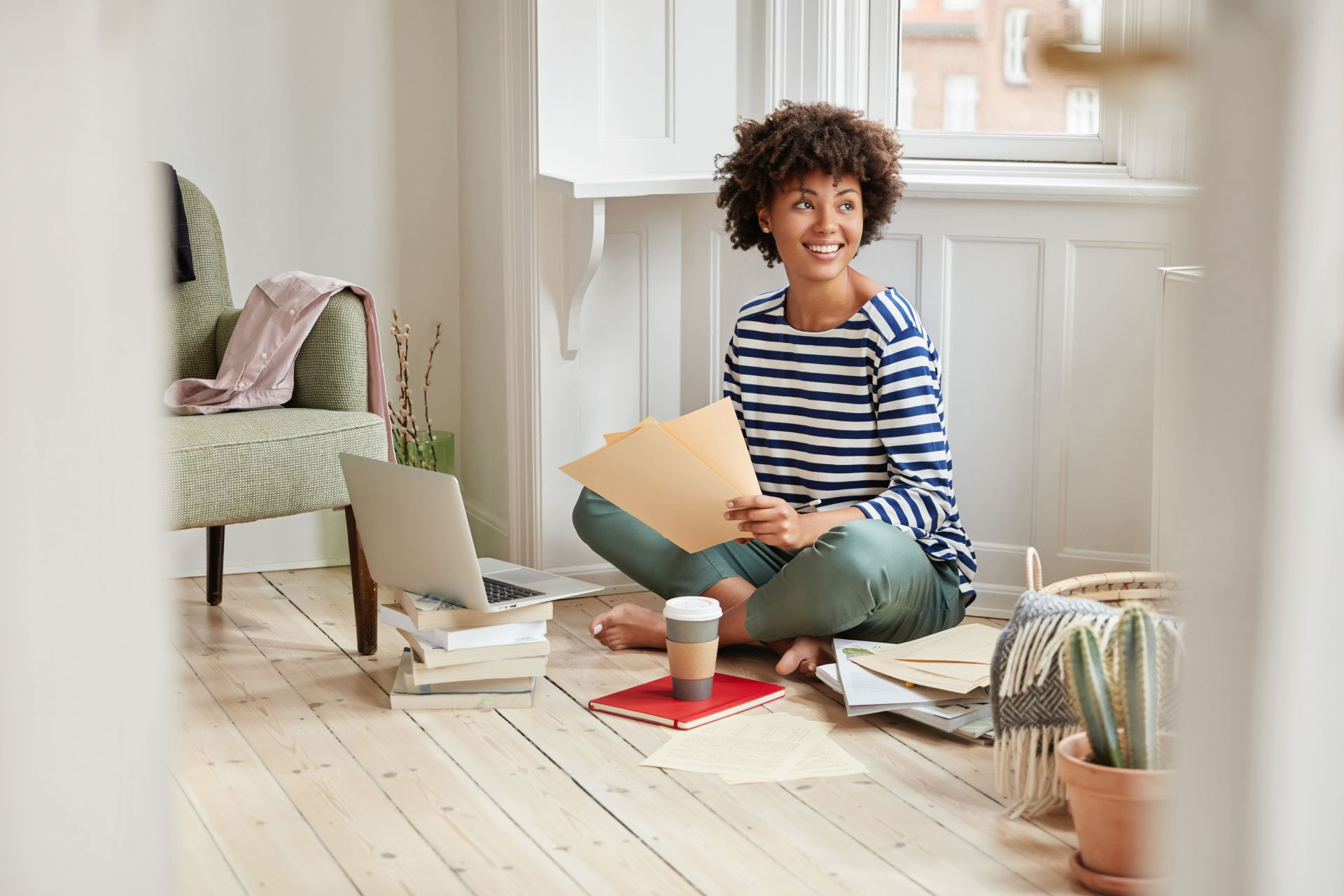 Frau sitzt auf dem Boden mit Laptop, Kaffee und Papieren, umgeben von Büchern in einem hellen Raum