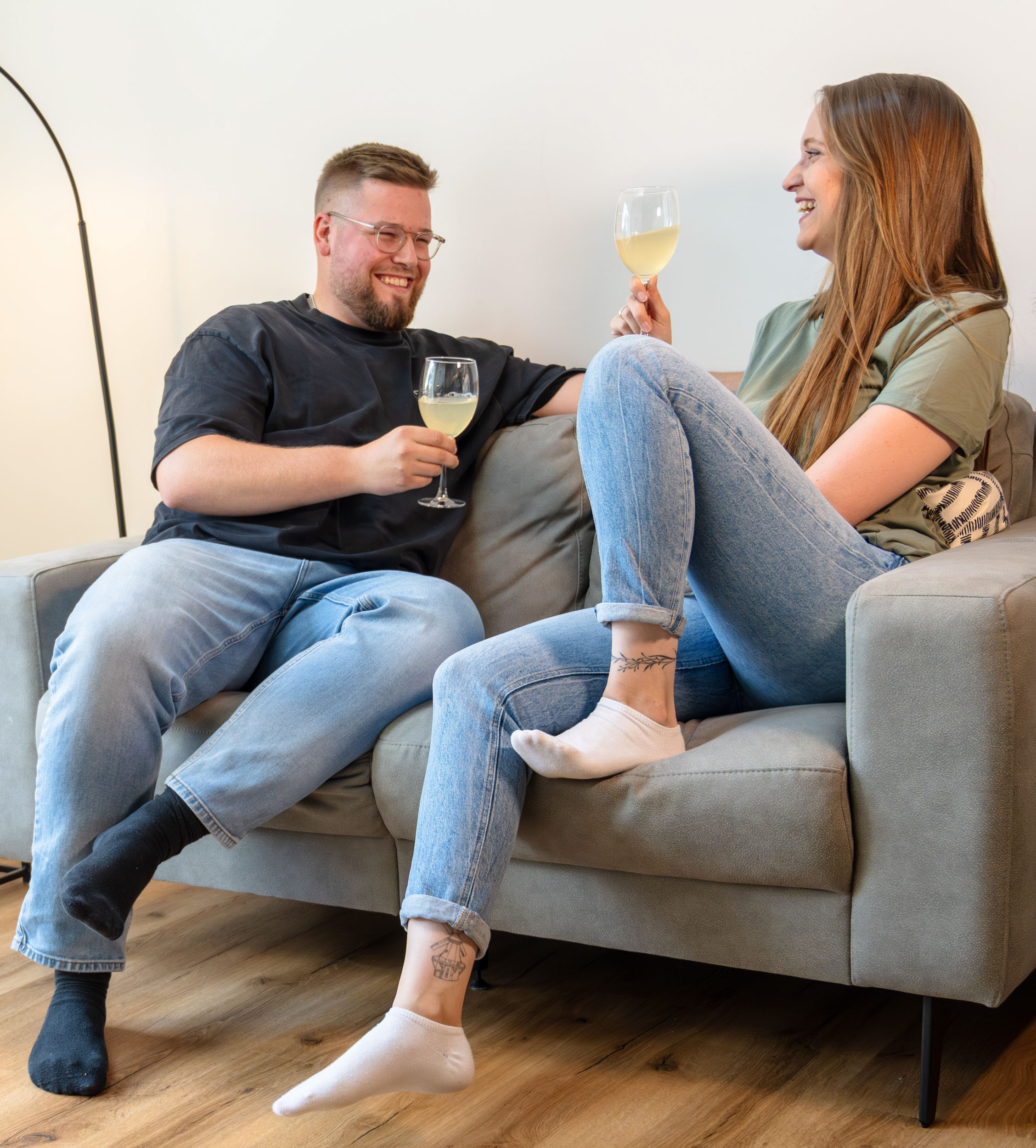 Junge Frau in Latzhose kniet auf dem Boden und hält lächelnd ein helles Laminatbrett in einem renovierungsbedürftigen Raum